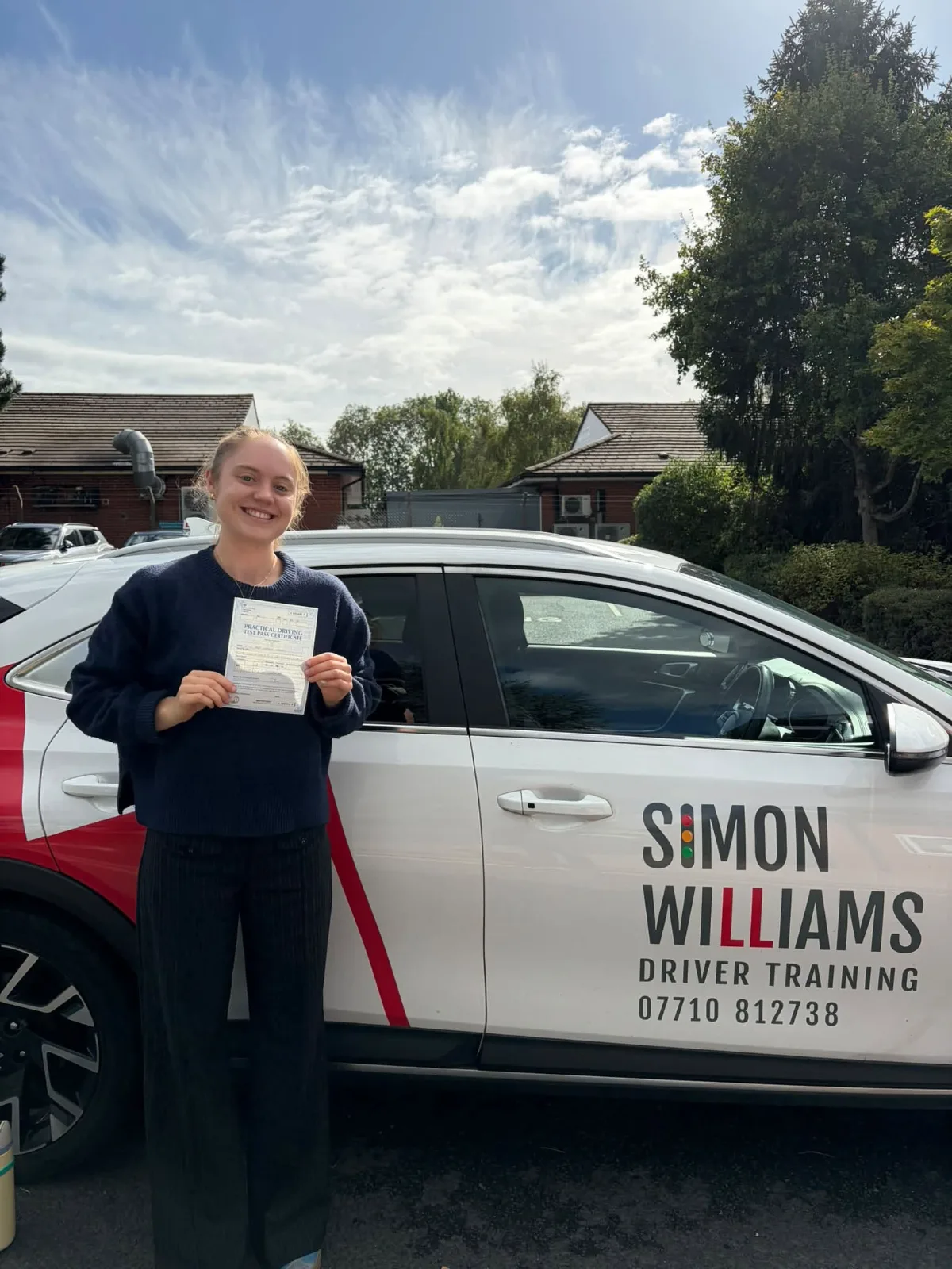 A smiling learner holding a pass certificate beside Simon Williams Driver Training's teaching car.