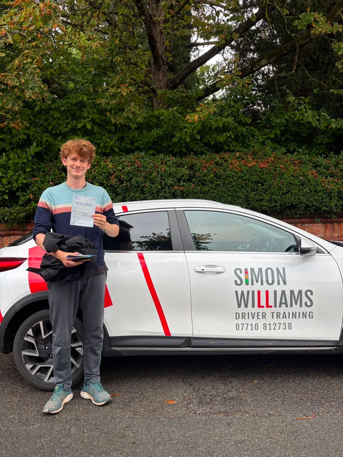 A learner in a striped jumper holding a pass certificate beside Simon Williams Driver Training's teaching car.