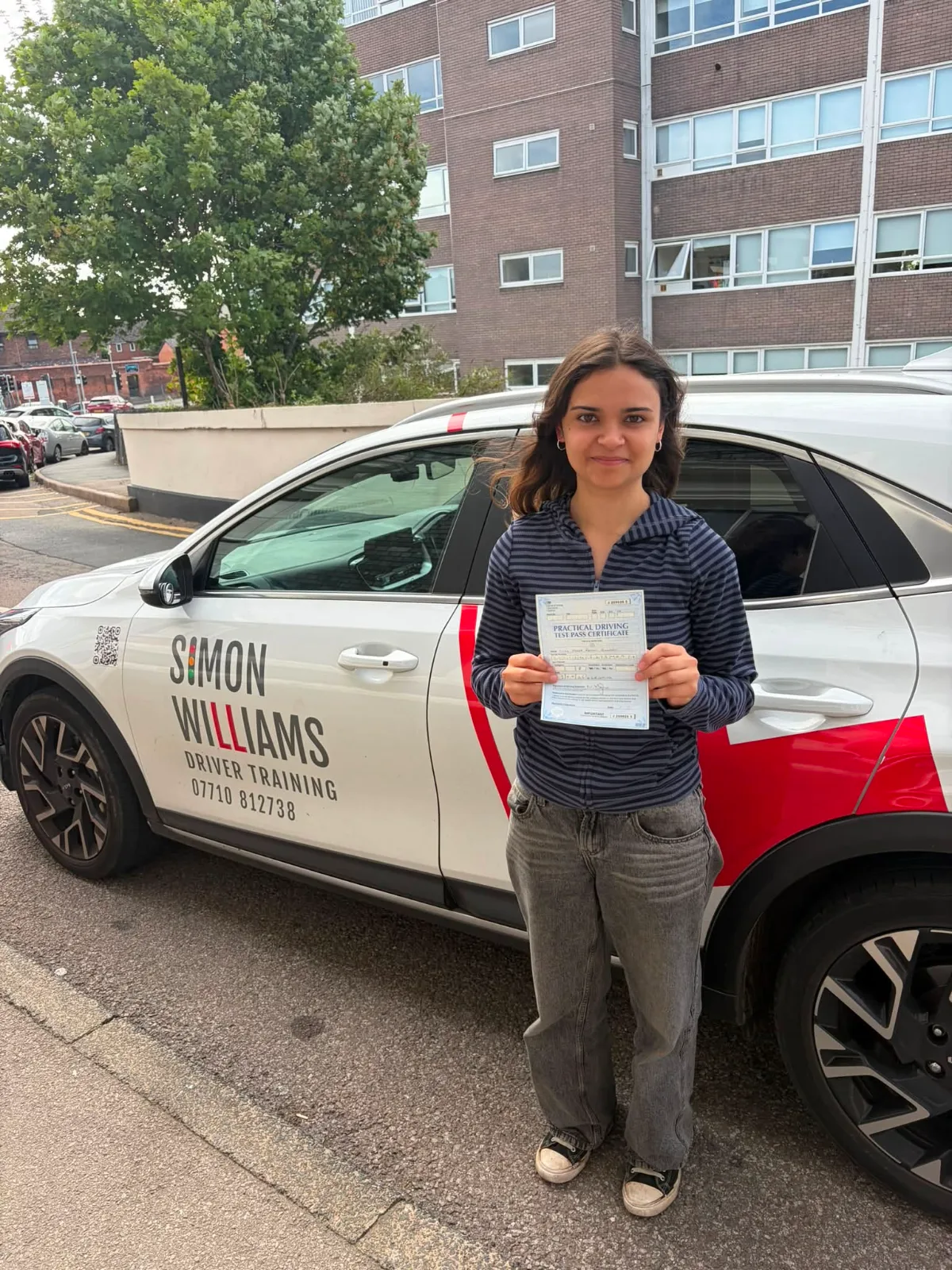 A learner in a striped top holding a pass certificate beside Simon Williams Driver Training's teaching car.