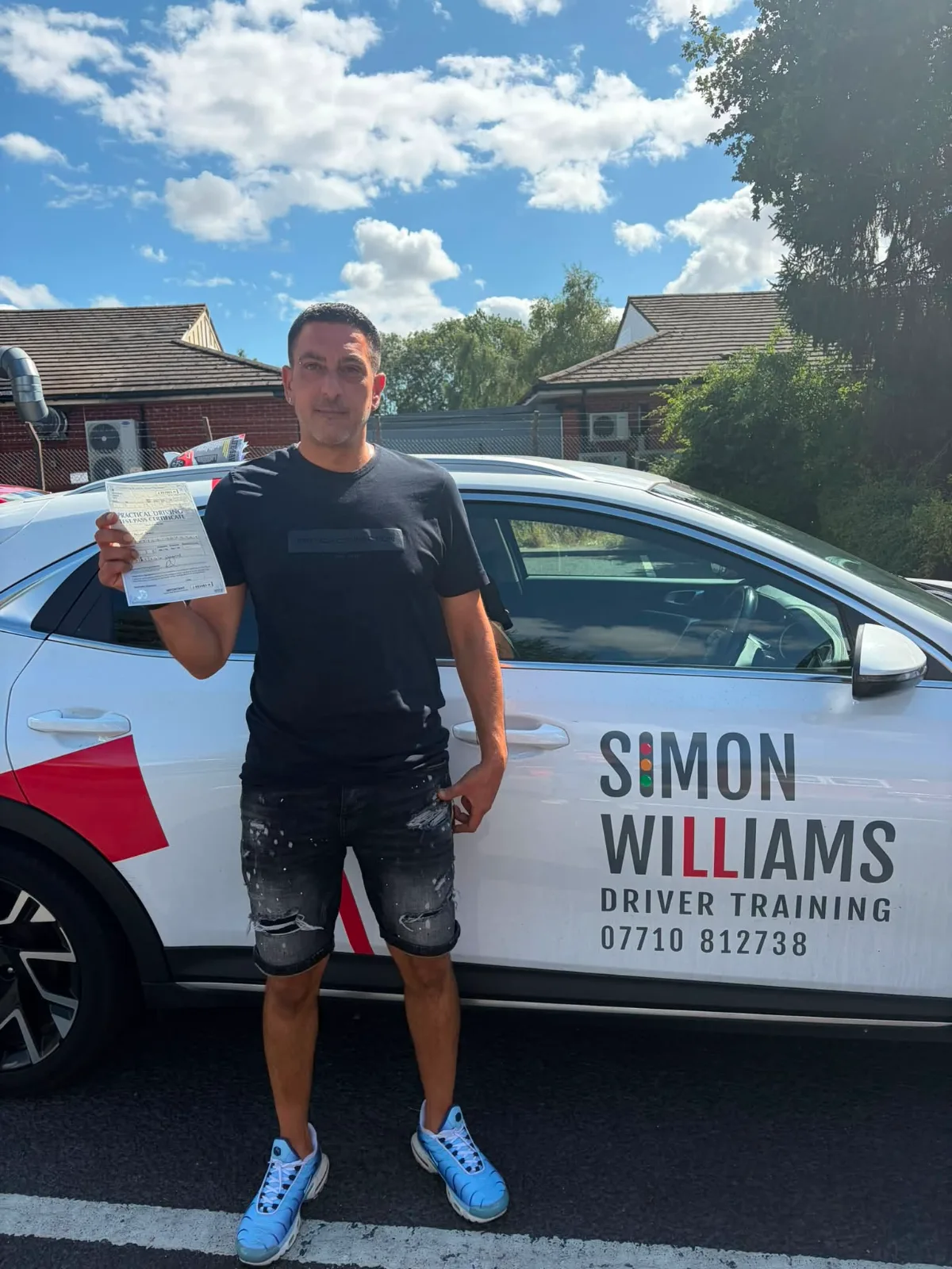 A learner in a black T-shirt holding a pass certificate beside Simon Williams Driver Training's teaching car.