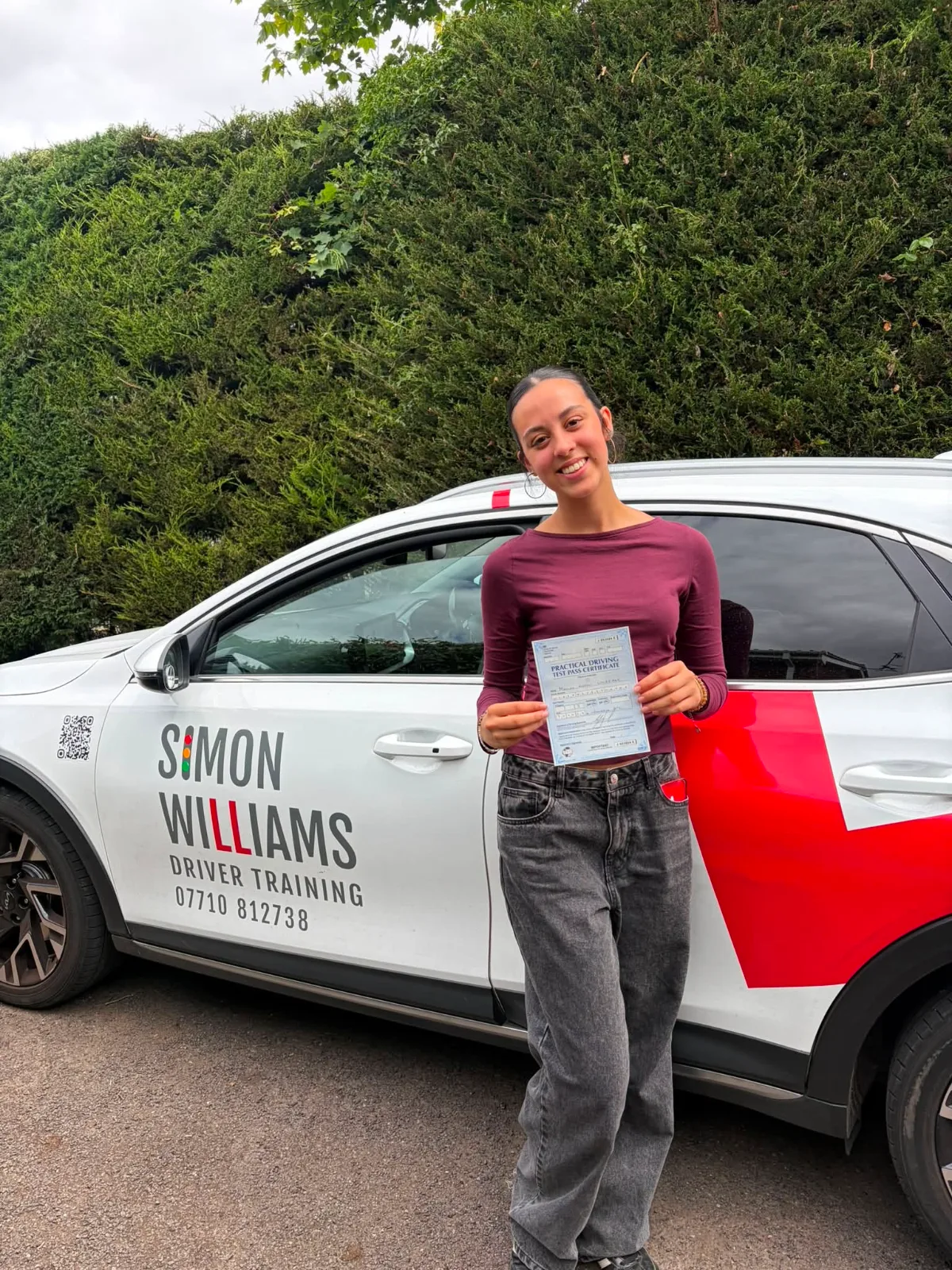 A smiling learner in a burgundy top holding a pass certificate beside Simon Williams Driver Training's teaching car.